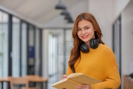 Photo for Female student smiling confidently holding copy book Standing at the school library Learn from home remotely Prepare homework online. - Royalty Free Image