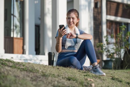 Photo for Outdoor shot of happy Asian woman exercising sitting on the grass in front of her after exercising. she looked satisfied, holding his smartphone to check exercise data.. - Royalty Free Image