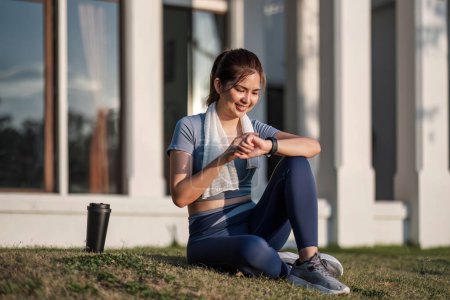 Téléchargez les photos : Plan extérieur de femme asiatique heureuse s'exerçant assis sur l'herbe devant elle après l'exercice. elle avait l'air satisfaite, tenant son smartphone pour vérifier les données d'exercice.. - en image libre de droit
