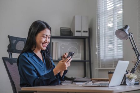 Photo for Happy Excited Asian young entrepreneur business woman using phone and laptop sitting on a desk at home workplace,. - Royalty Free Image