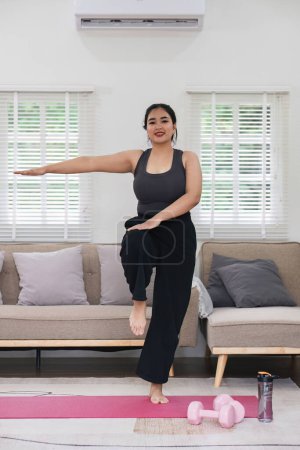 Téléchargez les photos : Femme asiatique en surpoids faisant de l'exercice à la maison sur un tapis de fitness. Belle femme caucasienne en vêtements décontractés Préparation à faire du yoga Exercice de pratique dans le salon. - en image libre de droit