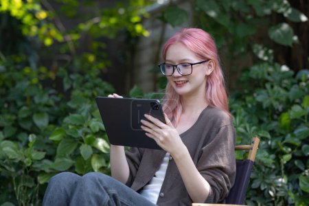 Téléchargez les photos : Femme asiatique enregistrer des données sur une tablette ou noter quelques idées tout en étant assis sur une chaise sous un arbre dans le parc - en image libre de droit