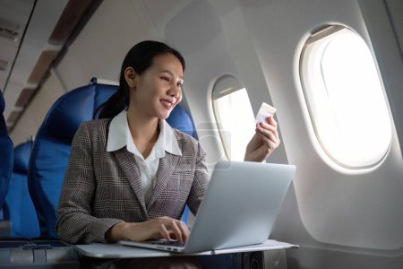 Téléchargez les photos : Attrayant passager asiatique femme dans l'avion assis dans un siège confortable à l'aide d'un ordinateur portable et carte de crédit, shopping en utilisant une connexion sans fil, voyager dans le style. - en image libre de droit