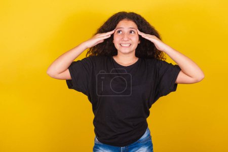 Foto de Brasileña, chica latinoamericana, para cabello afro, fondo amarillo, con mano en frente y cabeza, expresión de duda, olvido, vixi, cuestionamiento, arrepentimiento - Imagen libre de derechos