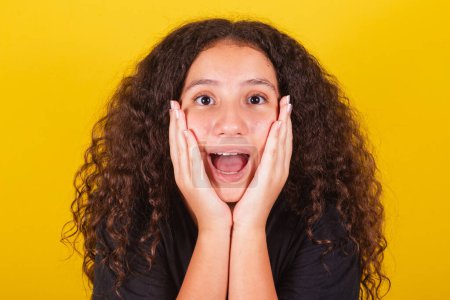 Photo for Brazilian, latin american girl, for, afro hair, yellow background, with hand on face, on chin, expression of surprise, surprised, happy, vibrant, joyful, scared, eyes wide. - Royalty Free Image