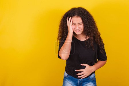 Photo for Brazilian, Latin American girl, for Afro hair, yellow background, Hands on belly and forehead, bellyache, abdominal discomfort, colic, diarrhea. headache, tiredness - Royalty Free Image