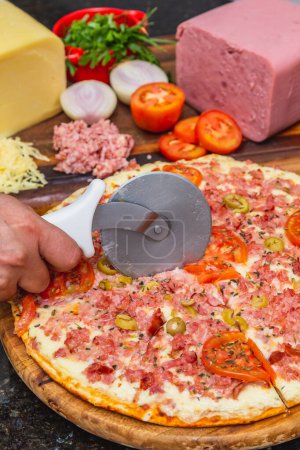Photo for Delicious mixed cheese and ham pizza being cut, ready, baked pizza, home pizza. next to wooden board with ingredients - Royalty Free Image