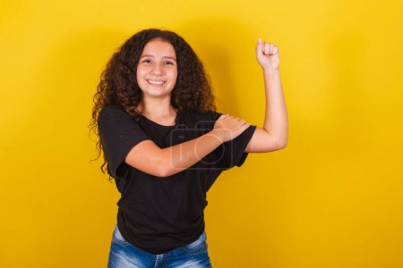 Téléchargez les photos : Brésilienne, Latino-américaine, pour cheveux afro, fond jaune, souriante, avec la main sur les biceps, heureuse, indiquant la force, l'autonomisation, le feliminisme, joyeuse, confiante, optimiste - en image libre de droit