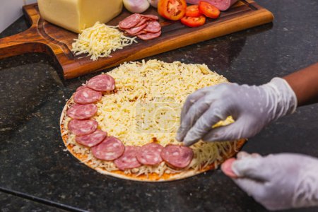 Foto de Preparación de pizza, esparcir anillos de pepperoni y rebanadas en masa de pizza, ingredientes frescos de pizza en tablero de madera. - Imagen libre de derechos
