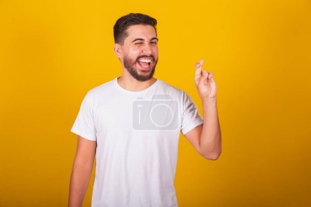 Foto de Hombre latinoamericano brasileño, dedos cruzados, signo de la suerte, animar, ansiedad, esperanzado, dibujar, feliz, alegre, fondo amarillo - Imagen libre de derechos