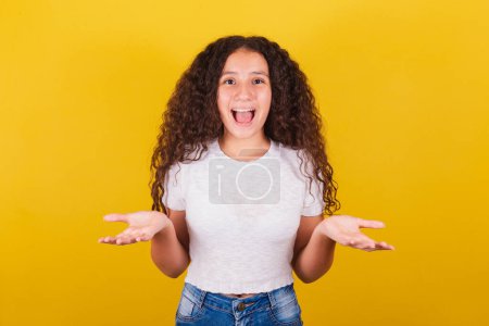 Foto de Chica brasileña latinoamericana para el pelo afro, fondo amarillo, sonriendo, haciendo expresión sorprendida con la cara y las manos, wow, sorprendida - Imagen libre de derechos
