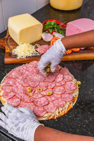 Foto de Preparación de pizza, esparcir aceitunas en pizza de pepperoni, ingredientes frescos de pizza en tablero de madera. - Imagen libre de derechos