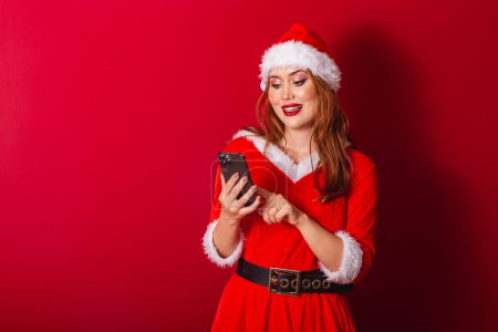 Téléchargez les photos : Belle femme rousse brésilienne, vêtue de vêtements de Noël, le Père Noël. tenant smartphone, tapant. - en image libre de droit