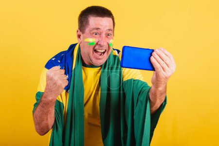 Téléchargez les photos : Homme adulte, fan de football brésilien, en utilisant drapeau, smartphone, regarder le jeu par téléphone portable. en ligne, streaming - en image libre de droit