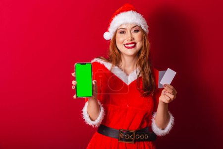 Photo for Beautiful Brazilian red-haired woman, dressed in Christmas clothes, Santa Claus. holding credit cards and smartphone with green screen in chroma. - Royalty Free Image