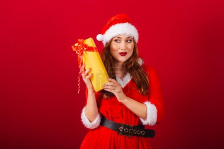 Photo for Beautiful Brazilian red-haired woman, dressed in Christmas clothes, Santa Claus. Doubt with what's in this yellow gift. - Royalty Free Image