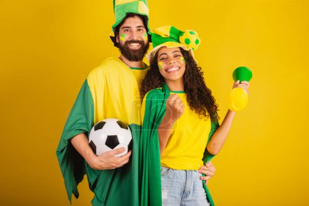 Foto de Pareja de aficionados al fútbol brasileño, vestidos con los colores de la nación, mujer negra, hombre caucásico. utilizando bandera y accesorios - Imagen libre de derechos