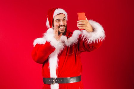 Téléchargez les photos : Homme brésilien, habillé en santa claus vêtements, tenant smartphone autoportrait avec téléphone portable, appel vidéo. - en image libre de droit