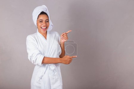 Photo for Beautiful Brazilian black woman, wearing bathrobe and towel. pointing with fingers, showing right, spa, aesthetics, delicacy. - Royalty Free Image