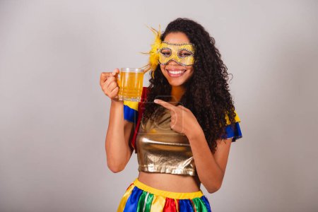 Téléchargez les photos : Belle femme brésilienne noire, avec des vêtements frevo, carnaval. dans le mascara, montrant un verre de bière, faire la fête dans le bar. - en image libre de droit