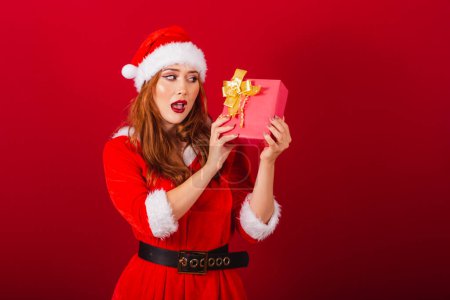 Photo for Beautiful Brazilian red-haired woman, dressed in Christmas clothes, Santa Claus. Doubts about what's in this red gift. - Royalty Free Image