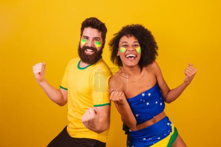 Téléchargez les photos : Couple de fans de football brésilien, habillé dans les couleurs du brésil, femme noire, homme caucasien. hurlant objectif - en image libre de droit