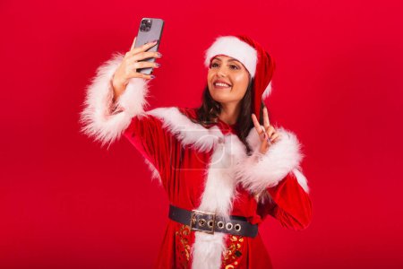 Photo for Brazilian woman dressed in Christmas clothes, Santa Claus. taking self portrait photo with smartphone. - Royalty Free Image