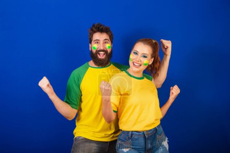 Téléchargez les photos : Couple caucasien, fans de football brésilien, tenant les bras dans la célébration - en image libre de droit
