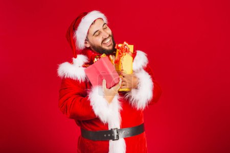 Foto de Hombre brasileño, vestido con ropa de Santa Claus, sosteniendo regalos. abrazando muy feliz - Imagen libre de derechos