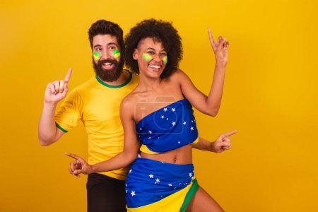Foto de Pareja de aficionados brasileños al fútbol, vestidos con los colores de Brasil, mujer negra, hombre caucásico. bailando juntos, divirtiéndose. - Imagen libre de derechos