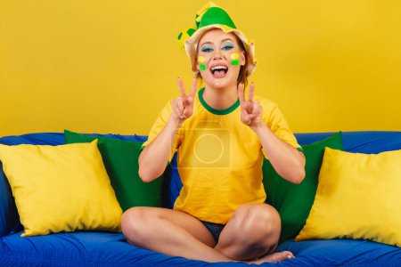 Photo for Caucasian woman, redhead, brazil soccer fan, brazilian, on the couch making peace and love symbol - Royalty Free Image