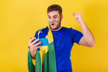 Téléchargez les photos : Homme caucasien brésilien, fan de football brésilien, célébrant objectif par téléphone portable, smartphone. Regarder par Streaming. - en image libre de droit