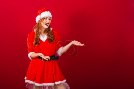 Téléchargez les photos : Belle femme rousse brésilienne, vêtue de vêtements de Noël, le Père Noël. montrer quelque chose à droite. - en image libre de droit