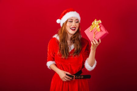 Foto de Hermosa brasileña pelirroja, vestida con ropa de Navidad, Santa Claus. Dudas sobre lo que hay en este regalo rojo. - Imagen libre de derechos