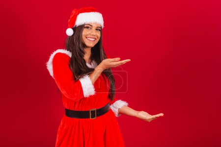 Téléchargez les photos : Belle Brésilienne vêtue de vêtements de Noël, le Père Noël. montrer quelque chose à droite. - en image libre de droit