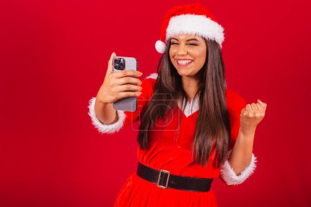 Téléchargez les photos : Belle Brésilienne vêtue de vêtements de Noël, le Père Noël. tenant smartphone célébrant. - en image libre de droit