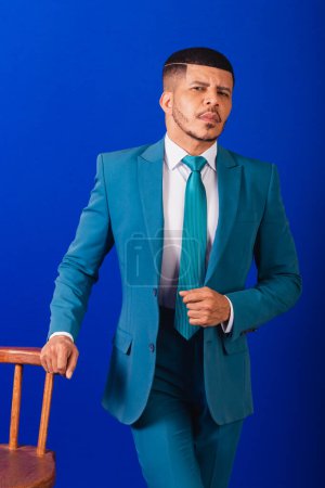 Téléchargez les photos : Homme noir brésilien, vêtu d'un costume et cravate bleue. homme d'affaires. pose de chaise en bois - en image libre de droit