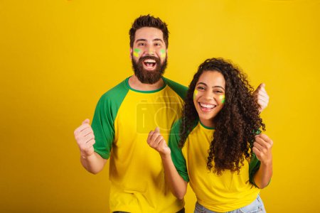 Téléchargez les photos : Couple de supporters brésiliens de football, vêtus aux couleurs de la nation, femme noire, homme caucasien. Torsion et vibration. - en image libre de droit