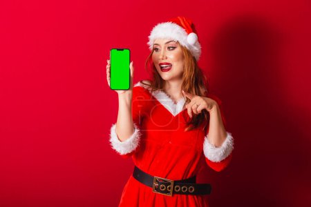 Foto de Hermosa brasileña pelirroja, vestida con ropa de Navidad, Santa Claus. mostrando smartphone con pantalla croma verde. - Imagen libre de derechos