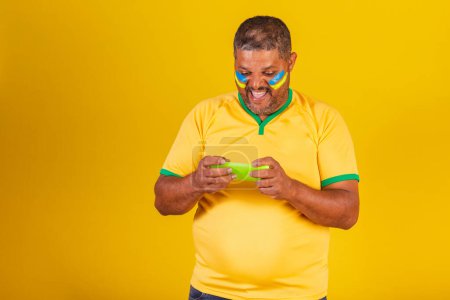 Foto de Hombre negro brasileño, fanático del fútbol de Brasil. viendo el juego en el móvil. smartphone - Imagen libre de derechos