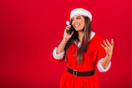 Photo for Beautiful Brazilian woman dressed in Christmas clothes, Santa Claus. holding smartphone voice call. - Royalty Free Image