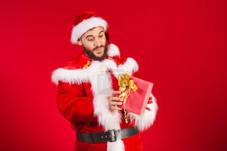 Photo for Brazilian man, dressed in santa claus clothes, holding red gift. - Royalty Free Image