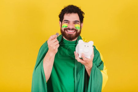 Photo for Man, brazilian, bearded, soccer fan from brazil, holding piggy bank and coin. concept of finance, economy, retirement. - Royalty Free Image