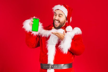 Photo for Brazilian man, dressed in santa claus clothes, holding smartphone with green chroma screen - Royalty Free Image