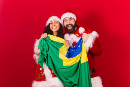 Photo for Brazilian couple, dressed for christmas, santa claus, mummy claus. holding brazil flag. black woman and caucasian man. - Royalty Free Image