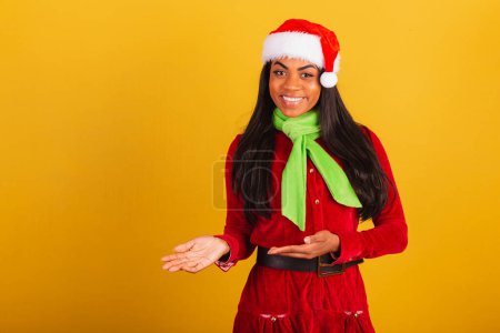 Photo for Beautiful black Brazilian woman, dressed in Christmas clothes, santa claus, presenting product or text on the side. left. - Royalty Free Image