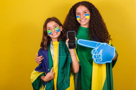 Photo for Sisters and friends cheerleaders from brazil, soccer fans, pointing to cellphone, internet, connected world, cellphone app. foam finger, world cup. Olympics. - Royalty Free Image