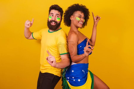 Photo for Couple of brazilian soccer fans, dressed in the colors of brazil, black woman, caucasian man. dancing together, having fun. - Royalty Free Image