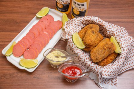 Photo for Chicken croquette and Cambu sausage,typical Brazilian snack, served with lemon slices, chili sauce and mayonnaise. On wooden table served with famous corona beer. - Royalty Free Image