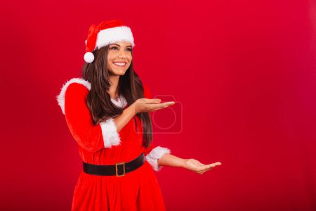 Photo for Beautiful Brazilian woman dressed in Christmas clothes, Santa Claus. showing something to the right. - Royalty Free Image